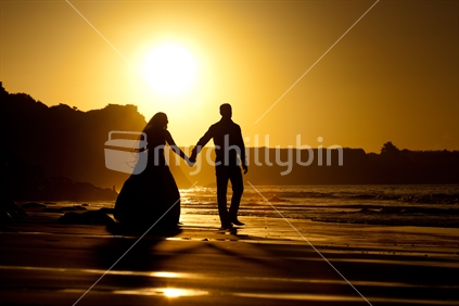 A bride & groom holding hands in front of a setting sun at the beach on their wedding day.