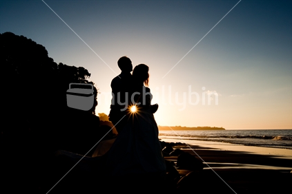 A bride & groom embracing in front of a setting sun at the beach on their wedding day.