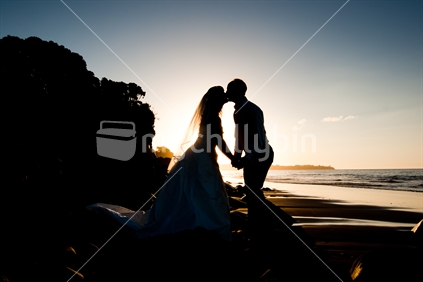 A bride & groom holding hands and kissing in front of a setting sun at the beach on their wedding day in New Zealand.
