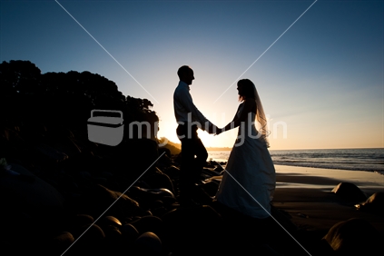 A bride & groom holding hands in front of a setting sun at a New Zealand beach on their wedding day.