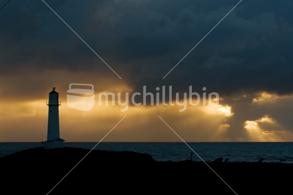 Cape Egmont lighthouse, Taranaki silhouetted by a setting sun through stormy clouds