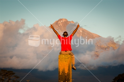 A man sitting on an old chimney in front of Mt Taranaki / Egmont in late afternoon sun.