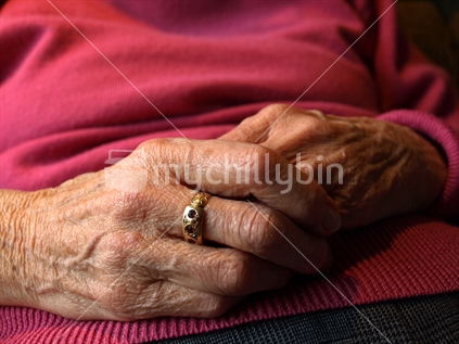 Older New Zealand woman sitting with hands on lap, and ring on finger. 