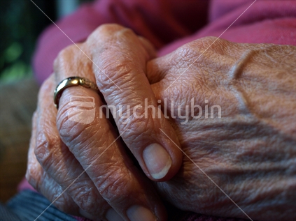 Older NZ woman sitting with hands on lap and ring on finger. 