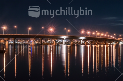 Night view of the Tauranga Harbour Bridge, its curving lines and lights and reflections.