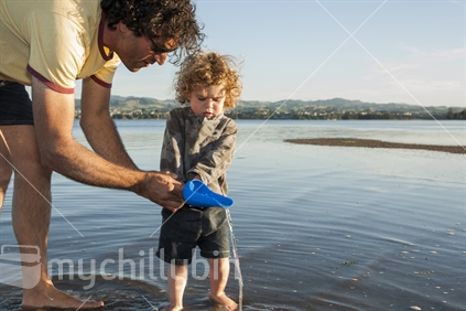 Father shows small son the wonders and physics of falling water as he pours water from a small beach spade, his son watches on trying to help.