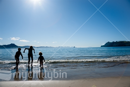Three children silhouetted and jumping small waves on water edge at beach (lens flare)