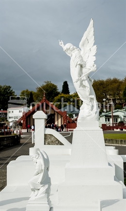 White stone angel in the Ohinemutu cemetery Rotorua. The marae wharenui in background.