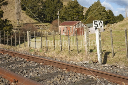 Railway and old cottage, Whangamomona. Perfect timeless bucolic pastoral scene with a traditionally deserted rural cottage illuminated by warm in the sunshine with  pasture on the hillside around and the rusty railway lines in foreground and 50 sign.