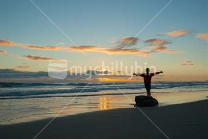 Boy balances on driftwood logs silhouetted by morning sunrise on beach.