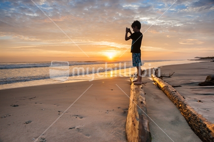 Boy balances on driftwood logs silhouetted by morning sunrise on beach, holding small device to take a photo.