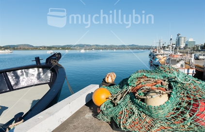 Fishing wharf, bow of boat and boats moored with nets piled in foreground, and city skyline, Tauranga.