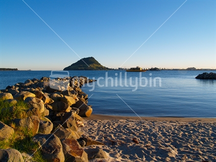 Mt Maunganui from Tauranga side of harbour, early morning on a clear day, with rocky foreground.