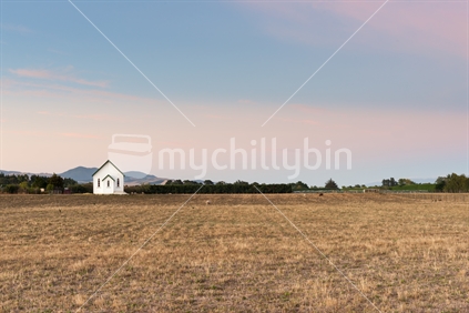 Small distant traditional wooden church stands alone in dry field at sunrise.