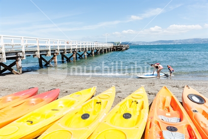 Summer day at Days Bay and wharf Wellington, New Zealand beach people at waters edge with paddle board with beach equipment yellow and orange kayaks and long wharf