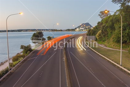 Highway red and yellow light streams along Takitimu Drive at dusk looking north with Mount Maunganui on horizon.