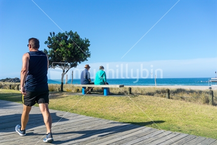 Eldery couple rest and look at scenic ocean view on bench seat along Mount Maunganui ocean-beach as man walks by on boardwalk.