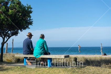 Elderly couple rest and look at scenic ocean view on becnh seat along Mount Maunganui ocean-beach while man waling on elevated rope appears to be walking on water beyond beach.