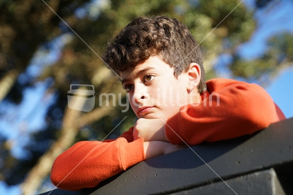 Boy looking ahead while resting on his arms top of wall with sun striking side of face.