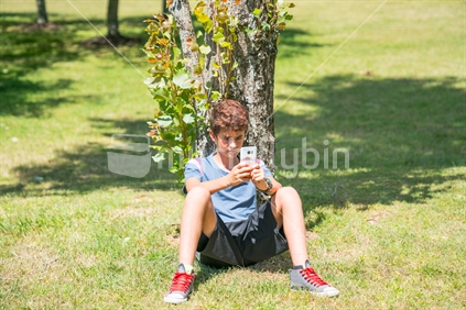 Boy sitting under tree on summer day in Cornwall Park using mobile phone with image of Mount Maunganui on cover.