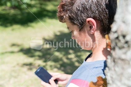 Boy leans on tree using mobile phone in selective focus on summer day in Cornwall Park.