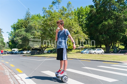 Boy licking ice cream riding hoverboard across pedestrian crossing on summer day in Cornwall Park.