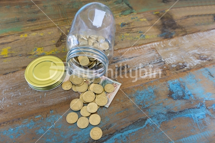 Savings jar with New Zealand currency coins and bank note spilling out on to wooden table - closeup