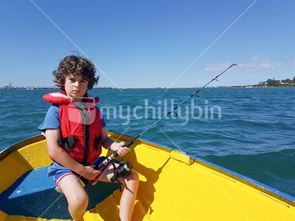 Kid with unruly hair fishing from small dinghy on Tauranga Harbour in bright yellow dinghy.
