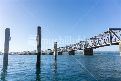 Tauranga harbour with steel railway bridge crossing with old piles projecting out of blue water.