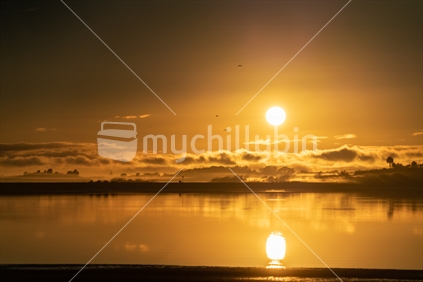 Golden glow of sunrise across bay on Tauranga harbour clouds and silhouette on misty horizon.