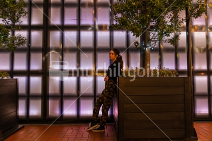 teenage girl leaning looking pensive on planter in front of panelled back-lit wall in Auckland's Britomart area waiting to meet someone.