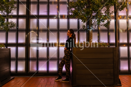Girl leaning looking pensive on planter in front of panelled back-lit wall in Auckland's Britomart area while she awaits arrival of friend