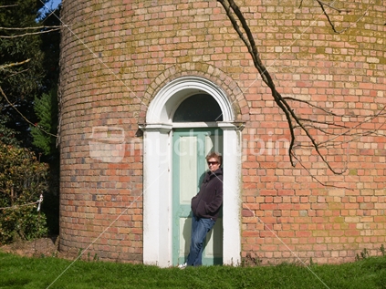 Woman sheltering from wind in doorway of Cambridge tall orange brick water tower on outskirts of town, New Zealand