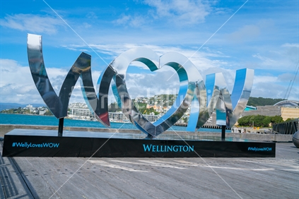 WELLINGTON, NEW ZEALAND - OCTOBER 1 2018; World of Wearable Art iconic shiny silver sign on city waterfront reflects city and arbour scenes and hillside of Oriental Bay background.