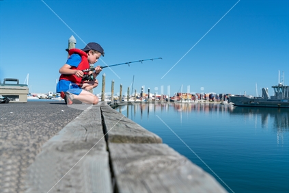 Boy sits on dock fishing wearing red life-jacket looking over edge into Tauranga harbour in marina New Zealand.