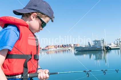 Boy stands on dock looking down into water fishing wearing red life-jacket and sunglasses Tauranga harbour in marina New Zealand.