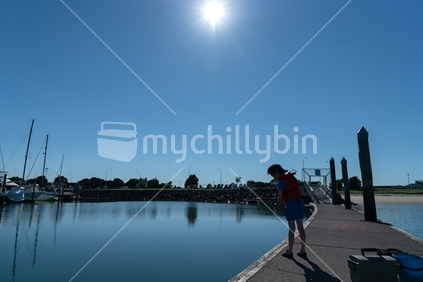 Small boy looking down  in red life jacket fishing from pier of marina on sunny calm morning silhouetted by morning sun
