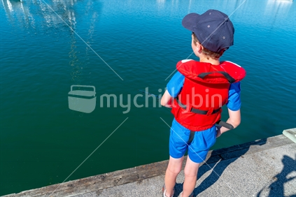Small boy from behind  in red life jacket fishing from pier of marina on sunny calm morning with moored boats and Tauranga harbour bridge in distance uner clear blue sky.