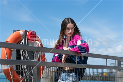 Attractive teenage woman stands looking deep in thought leaning on waterfront railing next to orange life buoy on Auckland waterfront, New Zealand.