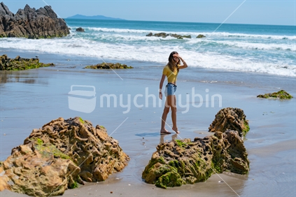 Young woman in bright yellow top walking near waters edge with rocky foreground at Mount Maunganui New Zealand.