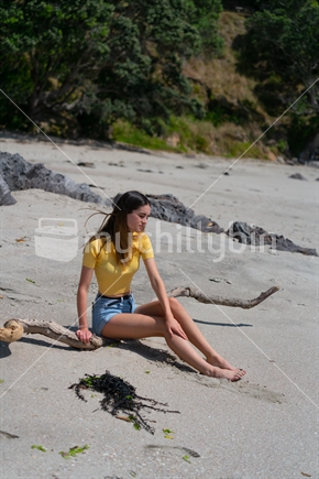 Teenage girl in yellow top and denim shorts sitting on piece of driftwood on beach at Mount Maunganui, New Zealand.