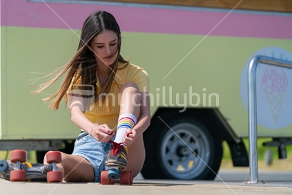 Teenage girl sits on pavement changing shoes for retro old pair roller skates with red laces beside retro caravan