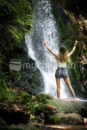 Shady low light inside NZ bush a young modern woman standing on rock looking at McLaren Falls waterfall with arms raisied, Tauranga New Zealand.