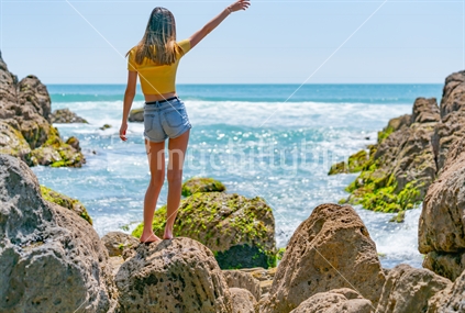 Girl in yellow top on coastal rocky seaside at Mount Maunganui, New Zealand standing on rocks with one arm raised.