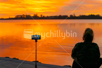 Tauranga New Zealand - April 25 2020; Brilliant sunrise being recorded on mobile device with silhouette of woman sitting by water's edge.