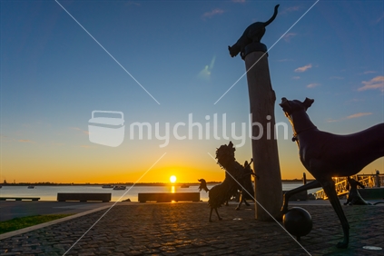 Hairy McClary characters of dogs and cat on pole silhouetted and back-lit by golden sunrise glow across Tauranga Harbour.