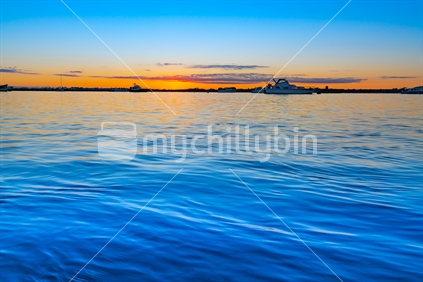 Sunrise over blue water of Tauranga harbour with intense golden glow on horizon.