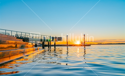 Sunrise over blue water of Tauranga harbour with intense golden glow on horizon with harbour steps and jumping platform.