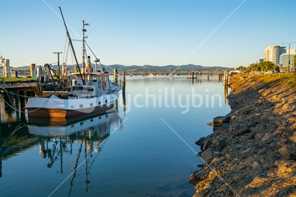 Waterfront at Fisherman's Wharf with moored commercial fishing boat and city downtown with railway bridge in distance in morning light at sunrise.