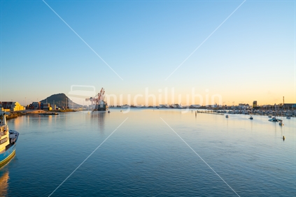 Tauranga harbour at sunrise with port facilities and landmark Mount Maunganui on golden horizon in distance.